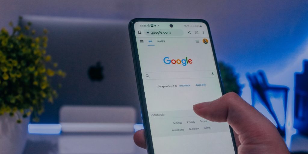 A person using Google search on a smartphone with a MacBook showing Apple logo placed in the background near the flower pot